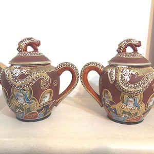 Op de afbeelding: Twee sierlijke, antieke theepotten met bijpassende deksels. De theepotten zijn dieprood met gouden en witte details, waaronder draakvormige tuiten en handvatten. Elke theepot heeft geschilderde figuren en ingewikkelde patronen.