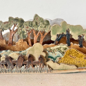 Peut inclure: Sculpture paysagère en céramique représentant des arbres, une clôture et un plan d'eau. Les arbres ont des feuilles et des troncs verts et bruns. La clôture est marron et l'eau est bleue et verte. La sculpture est peinte dans des tons terre.