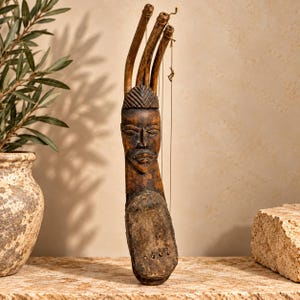 Könnte beinhalten: Ein dunkelbraunes, geschnitztes Musikinstrument aus Holz mit einem skulptierten menschlichen Gesicht an der Oberseite, drei nach oben gebogenen Holzhälsen und dünnen Saiten, die an einer abgerundeten, ausgehöhlten Basis mit vier Löchern befestigt sind.
