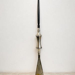 Op de afbeelding: Een antieke schoenlepel met een lange, zwarte handgreep en een gebogen haak aan de bovenkant. Het centrale gedeelte is zilverkleurig en de schoenlepel zelf heeft een versleten, metallic kleur. Het item wordt gepresenteerd tegen een effen witte achtergrond.