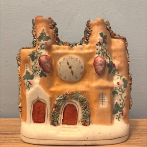 Puede incluir: Un reloj de cerámica con forma de edificio, con una esfera central. El edificio está pintado en tonos naranja y blanco, con puertas rojas y vides decorativas. El reloj tiene un aspecto envejecido y antiguo.