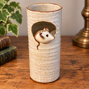 May include: A cylindrical ceramic vase with a speckled off-white glaze and horizontal ribbed texture. A sculpted mouse head emerges from a circular cutout near the top of the vessel. The rim and the mouse details feature a brown accent colour.