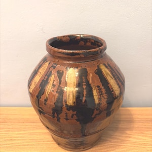 Könnte beinhalten: Eine handgefertigte, runde Keramikvase mit breiter Öffnung. Die Vase hat einen braunen Sockel mit vertikalen Streifen in Schwarz und Beige. Der Innenraum ist dunkelbraun, und die Vase steht auf einem Holzregal.