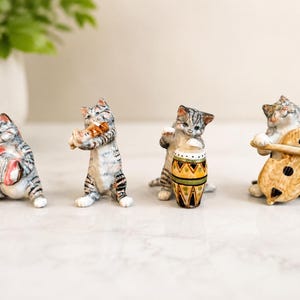 Può includere: Un set di quattro statuette di gatti in ceramica, dipinte con motivi tigrati grigi. I gatti sono raffigurati come musicisti: uno suona una fisarmonica rossa e bianca, uno un flauto, uno un tamburo giallo e verde e uno un contrabbasso marrone chiaro.