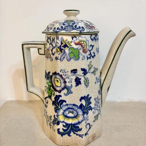 Può includere: Una caffettiera antica in ceramica con coperchio. La caffettiera presenta una base color crema con un motivo floreale blu, giallo, verde e rosso. Il manico e il beccuccio sono rifiniti con bordi verdi. La caffettiera ha una glassa craquelé.