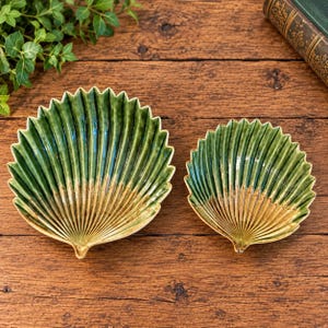 Può includere: Due ciotole in ceramica a forma di conchiglia di capasanta, caratterizzate da una finitura lucida con una sfumatura che va dal verde bosco intenso sui bordi a un caldo color sabbia alla base, con evidenti scanalature verticali radiali.