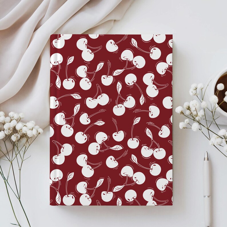 May include: A rectangular notebook with a burgundy cover featuring a repeating pattern of white cherries. The cherries have stems and leaves. The notebook is on a white surface with a pen, a cup of coffee, and white flowers.