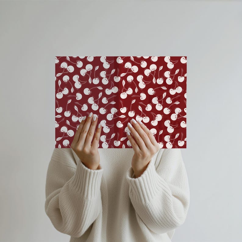 May include: A rectangular sheet with a repeating pattern of white cherries on a deep red background. The sheet is held up by a person wearing a white sweater.