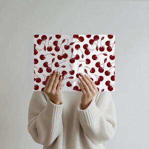 May include: A white book cover with a repeating pattern of dark red cherries. The cherries have stems and leaves. The book is held by a person wearing a white sweater.