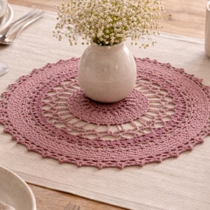 May include: A dining table setting featuring a mauve crocheted doily with a floral design, a small white vase with white flowers, and a glass of water. Silverware and plates are also visible, creating a soft, inviting ambiance.