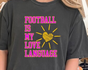 May include: Dark gray t-shirt with the text "FOOTBALL IS MY LOVE LANGUAGE" in pink with a yellow heart and sun design. The shirt is part of a football-themed collection, perfect for game day.