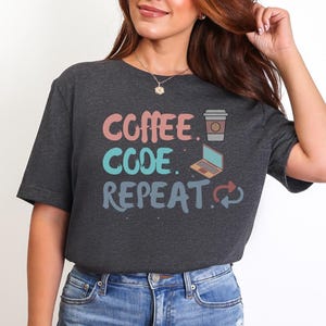 May include: Dark gray t-shirt with the words "COFFEE CODE REPEAT" in a colorful design. The design includes a coffee cup, a laptop, and a recycling symbol. The shirt is made of a soft material and is suitable for casual wear.