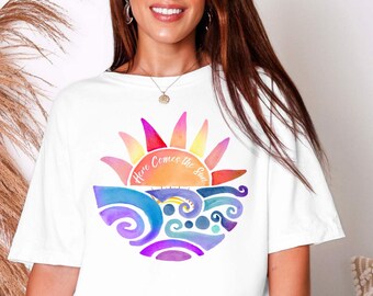 May include: White t-shirt featuring a watercolor design of a colorful sun and ocean waves. The text "Here Comes the Sun" is arched across the sun. The design includes shades of pink, orange, yellow, blue, and purple.