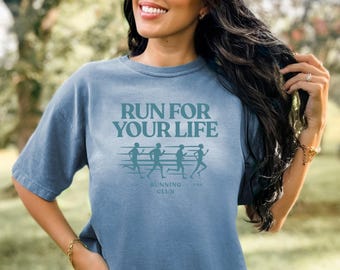 May include: A blue t-shirt with the text "RUN FOR YOUR LIFE" in a teal color. Below the text are four running figures and the words "RUNNING CLUB USA". The shirt is a solid color and has short sleeves.