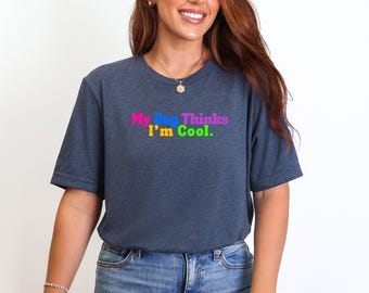 May include: A navy blue t-shirt with the text "My Dog Thinks I'm Cool" in colorful, rainbow-like lettering. The shirt is a casual style, suitable for everyday wear.