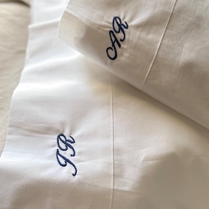 May include: White pillowcases with navy blue embroidered initials. One pillowcase features the initials "AR", the other "JR". The folded pillowcases are stacked, highlighting the clean fabric and personalised embroidery.