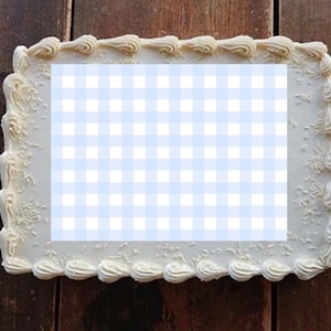 May include: A rectangular cake with white frosting and decorative swirls around the edges. The center features a light blue and white gingham pattern. The cake sits on a dark wooden surface.