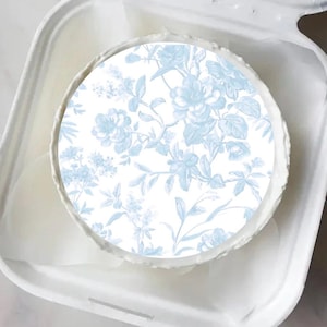 May include: A round, white object with a light blue floral design, possibly a cake or dessert, sits inside a white container. The floral pattern features roses and other flowers. The container is open, revealing the object within.