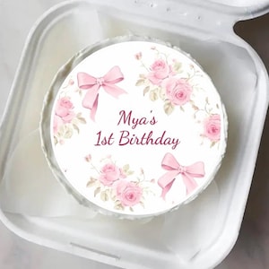 May include: A round cake with a floral design and the text "Mya's 1st Birthday" in a white container. The cake features pink roses, bows, and leaves. The container is white.