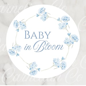 May include: A round white sticker with the text "BABY in Bloom" in a soft blue script. The text is surrounded by a delicate wreath of light blue watercolor flowers and green stems, perfect for baby shower decorations.