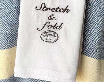 May include: White kitchen towel with the words "Stretch & Fold" embroidered in brown script, along with a bread illustration. The towel is hanging over a blue and white patterned fabric.