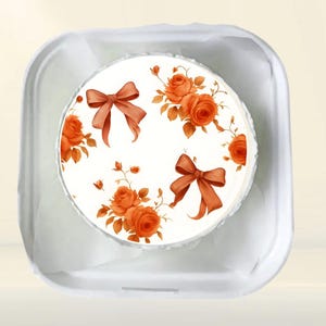 May include: A round cake with a white frosting base, decorated with orange roses and brown bows. The cake is in a white container with a light beige background.