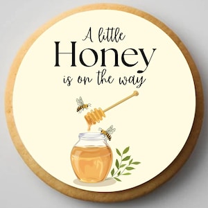 May include: A round cookie with a light yellow center and a brown edge. The center features a jar of honey, a honey dipper, and two bees. The text "A little Honey is on the way" is written in black script.