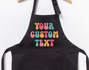 May include: Black apron with a pocket and the text "YOUR CUSTOM TEXT" in colorful, retro-style lettering. The apron hangs from a wooden hanger against a white background.