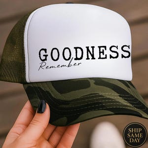 Può includere: Cappellino da camionista con un pannello frontale bianco con la scritta "GOODNESS" sopra "Remember" in corsivo nero. La visiera e la parte inferiore del cappello sono mimetiche e il retro in rete è verde oliva.