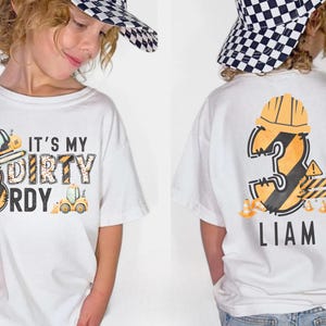 Peut inclure: T-shirt blanc avec un motif sur le thème de la construction. Le devant affiche le texte "It's My Dirty 3rd B-Day" avec des graphiques de véhicules de chantier. Le dos montre un grand chiffre 3 avec un casque et le nom "Liam".