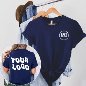 May include: Navy blue t-shirt with rolled sleeves and a white circular logo that says "YOUR LOGO". The back of the shirt also has the words "YOUR LOGO" in white. A brown hat and denim shirt hang in the background.