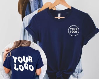 May include: Navy blue t-shirt with rolled sleeves and a white circular logo that says "YOUR LOGO". The back of the shirt also has the words "YOUR LOGO" in white. A brown hat and denim shirt hang in the background.