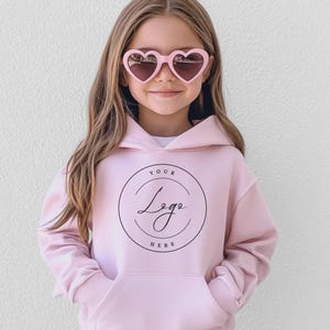May include: A young person wearing a light pink hoodie with a front pocket and a hood. The hoodie features a circular logo with the text "Your Logo Here". The person is also wearing pink heart-shaped sunglasses.