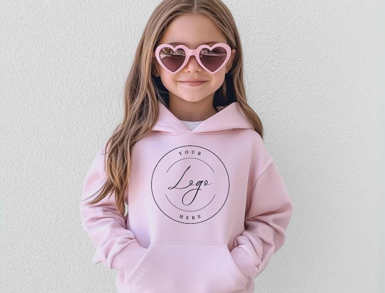 May include: A young person wearing a light pink hoodie with a front pocket and a hood. The hoodie features a circular logo with the text "Your Logo Here". The person is also wearing pink heart-shaped sunglasses.