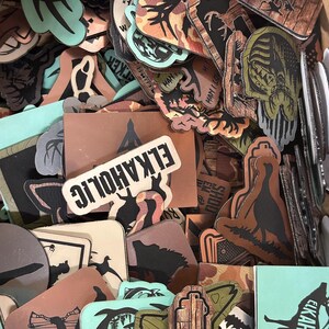 May include: A selection of rectangular and uniquely shaped patches. The patches feature outdoor-themed designs, including elk, trees, and camouflage patterns. Some patches have the text "ELKAHOLIC" and "MAN I LOVE HUNTING". The colour palette includes shades of brown, green, and teal.