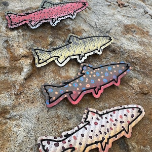 Puede incluir: Cuatro adornos con forma de pez con contornos negros y diversos patrones. Uno es rosa con puntos negros, otro es amarillo con un patrón de escamas, un tercero es gris con puntos azules y rosas, y el último es blanco con puntos marrones y rojos.
