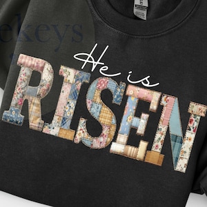 May include: Black sweatshirt featuring the text "He is RISEN" in a patchwork design. The letters are crafted from fabric with floral and geometric patterns. An Easter egg is visible in the upper right corner.