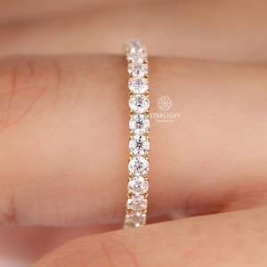 May include: A gold band ring featuring a continuous row of round, clear gemstones. The ring is set with multiple small, sparkling stones. The ring is shown on a person's finger.
