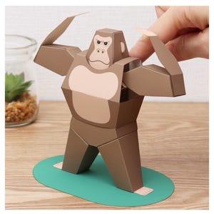 Op de afbeelding: Een bruine papieren gorilla-sculptuur met een lichtbruin gezicht en borst, staand op een groene ovale basis. De armen van de gorilla zijn omhoog, en de sculptuur is gemaakt van gevouwen papier. Een leuk, decoratief item.