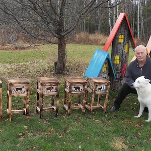 May include: A collection of rustic wooden stools with wildlife art. Each stool features a different animal silhouette, including a moose, bear, and wolf. The stools are displayed outdoors on a grassy lawn.