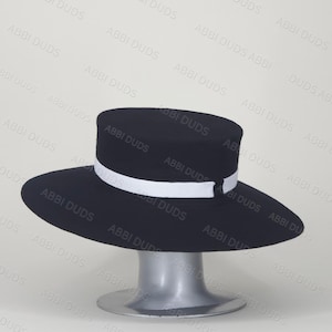 Pu&ograve; includere: Un cappello nero con corona piatta e tesa larga leggermente inclinata verso il basso. Una fascia bianca circonda la base della corona, presentando un piccolo dettaglio sovrapposto sul davanti. Il cappello &egrave; esposto su un supporto argentato liscio.