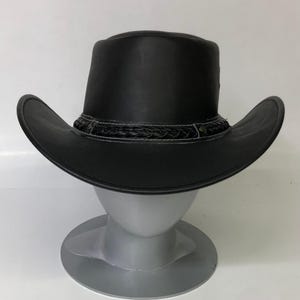 May include: A black leather cowboy hat with a wide brim and a braided band around the crown. The hat is displayed on a gray mannequin head. The hat's brim is curved, and the hat has a classic Western style.