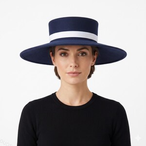 May include: A navy blue flat-top hat with a wide brim and a solid white band encircling the base of the crown. The hat features a structured, smooth finish and a clean, circular silhouette that sits horizontally across the head.