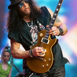 Puede incluir: Un músico con sombrero de copa negro con banda plateada, gafas de sol oscuras y camiseta negra con gráfico, tocando una guitarra eléctrica Gibson de color dorado. La guitarra presenta un cuerpo con corte y botones de control dorados.
