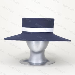 Pu&ograve; includere: Un cappello blu navy a corona piatta con tesa larga e una fascia bianca tinta unita che circonda la base della corona. Il cappello &egrave; esposto su un manichino a forma di testa color argento su uno sfondo chiaro con il testo ABBI DUDS.