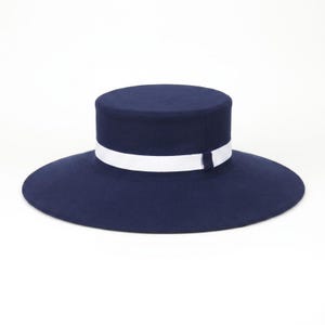 Pu&ograve; includere: Un cappello blu navy a corona piatta caratterizzato da un'ampia tesa circolare e da un nastro bianco nitido avvolto attorno alla base della corona. Il cappello presenta una finitura strutturata e liscia e una silhouette pulita e simmetrica.