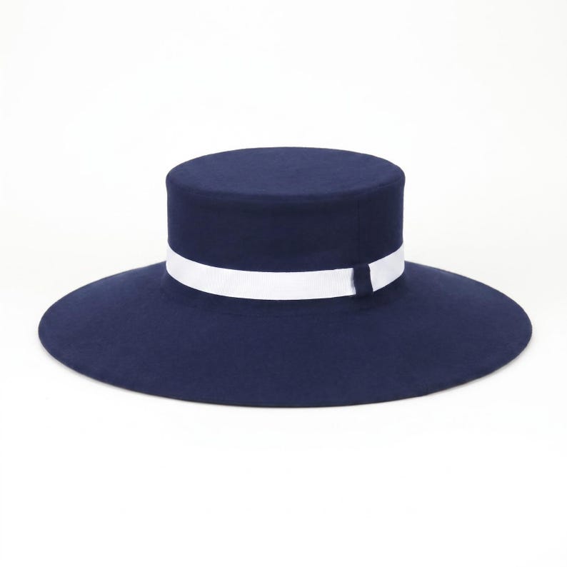 Pu&ograve; includere: Un cappello blu navy a corona piatta caratterizzato da un'ampia tesa circolare e da un nastro bianco nitido avvolto attorno alla base della corona. Il cappello presenta una finitura strutturata e liscia e una silhouette pulita e simmetrica.