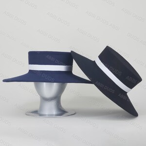 Pu&ograve; includere: Due cappelli blu navy a tesa larga con corona piatta e nastro bianco. Un cappello &egrave; posizionato su un supporto per testa di manichino argentato, mentre il secondo &egrave; inclinato accanto, mostrando la struttura della tesa e della corona.