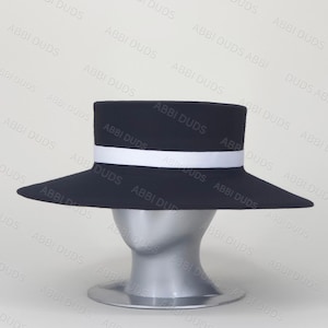 Pu&ograve; includere: Un cappello nero a tesa piatta con una tesa larga e una fascia bianca solida che circonda la base della corona. Il cappello &egrave; esposto su una testa di manichino color argento. Il testo ABBI DUDS &egrave; ripetuto come filigrana sull'immagine.