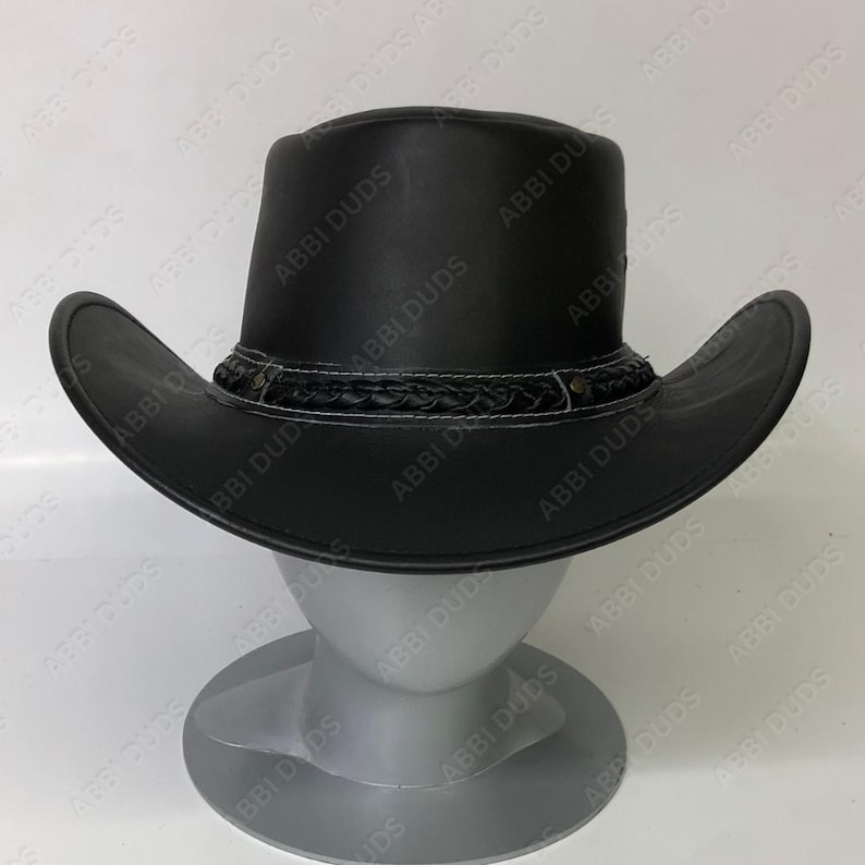 May include: A black leather cowboy hat with a wide brim and a decorative band around the crown. The hat is displayed on a mannequin head. The band features a braided design with white stitching and small metal studs. The hat's shape is classic, with a slightly curved brim.